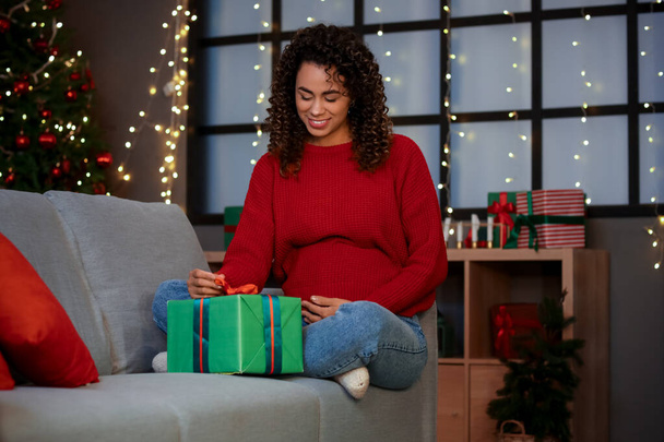 Femme afro-américaine enceinte avec cadeau de Noël assis à la maison - Photo, image