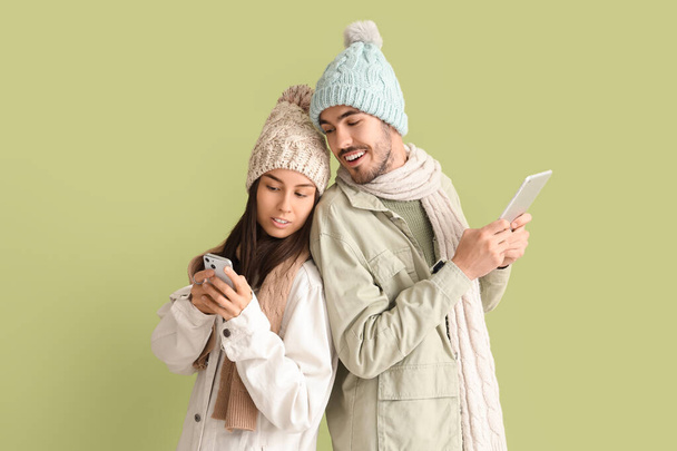 Szerető pár téli ruhák segítségével mobiltelefon és tabletta számítógép zöld alapon - Fotó, kép