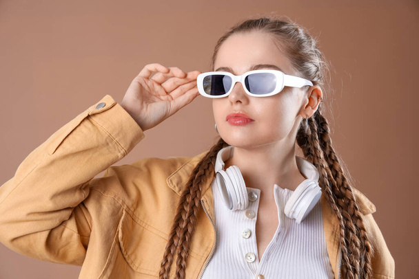 Bella giovane donna con occhiali da sole e cuffie su sfondo marrone - Foto, immagini