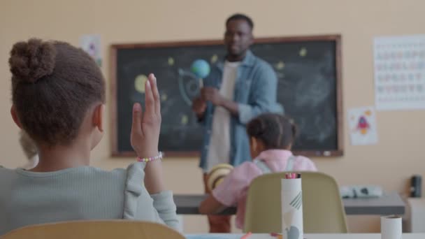 Selectieve focusshot van het kleine Afro-Amerikaanse meisje aan het bureau in de klas, hand opsteken en vragen stellen tijdens astronomieles op de basisschool - Video