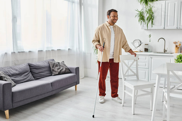 cheerful indian man with visual impairment in casual attire using walking stick while at home - Photo, Image