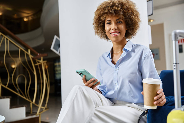 krullend en gelukkig Afrikaans amerikaanse vrouw met koffie te gaan en smartphone wachten in hotel lobby - Foto, afbeelding