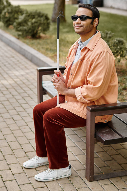 homme aveugle indien joyeux en veste orange assis à l'extérieur sur le banc avec bâton de marche et des lunettes - Photo, image