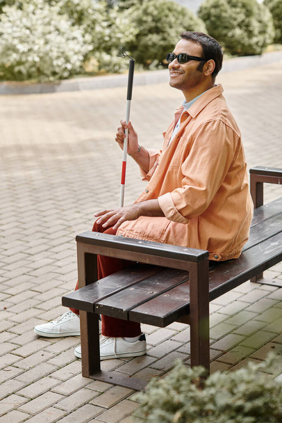 joyous indian blind man in orange jacket sitting outside on bench with walking stick and glasses - Photo, Image