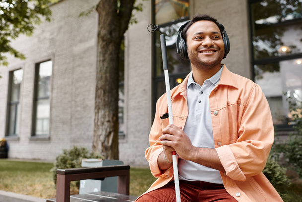 vrolijke indiaanse man met blindheid in hoofdtelefoon met wandelstok genieten van muziek terwijl buiten - Foto, afbeelding
