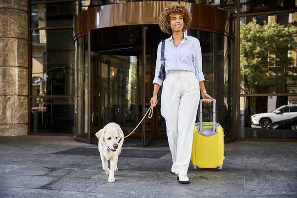 Femme afro-américaine aux cheveux bouclés avec son chien et ses bagages sortant de l'hôtel acceptant les animaux de compagnie - Photo, image