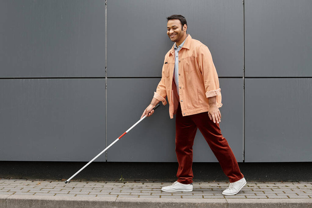 vrolijke indiaanse blinde man in oranje jas met helpende stok lopen met grijze muur op de achtergrond - Foto, afbeelding
