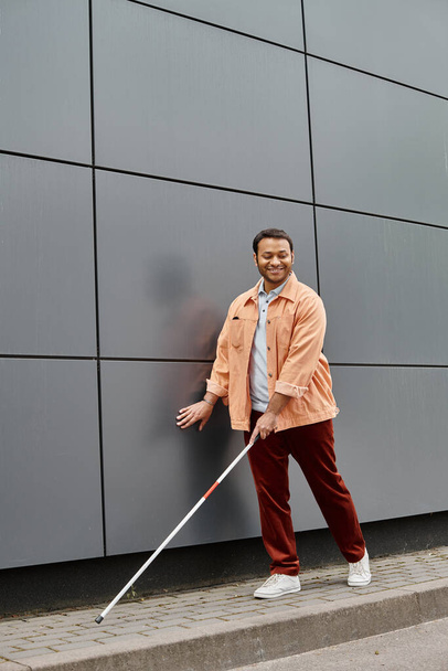 alegre indio ciego hombre en naranja chaqueta con ayuda palo caminando con gris pared en telón de fondo - Foto, imagen
