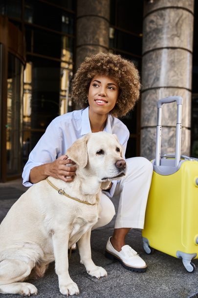 Femme afro-américaine souriante câlinant labrador près des bagages devant un hôtel acceptant les animaux de compagnie - Photo, image