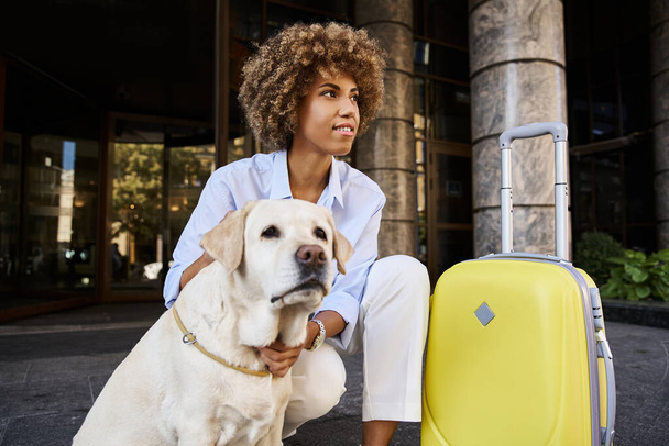 alegre mujer afroamericana acariciando labrador cerca de equipaje fuera de un hotel que acepta mascotas - Foto, Imagen
