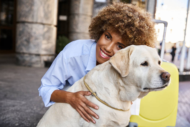 Smiling african american woman cuddling labrador near yellow luggage outside a pet-friendly hotel - Photo, Image