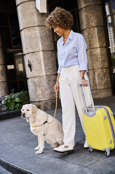 ペットフレンドリーなホテルの入り口近くに立っている犬と荷物を持つ肯定的なアフリカ系アメリカ人女性 - 写真・画像