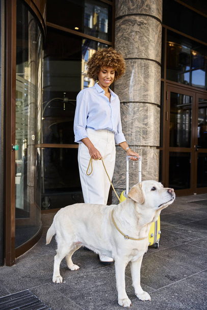 eccitata donna afroamericana con cane e bagagli in piedi vicino all'ingresso dell'hotel pet friendly - Foto, immagini