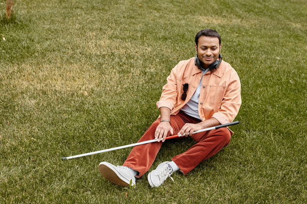 alegre indio ciego hombre de chaqueta naranja sentado en la hierba con auriculares y bastón - Foto, imagen