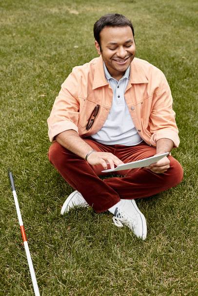 fröhlicher indischer Mann mit Blindheit in orangefarbener Jacke sitzt auf Gras und liest Blindenschrift - Foto, Bild