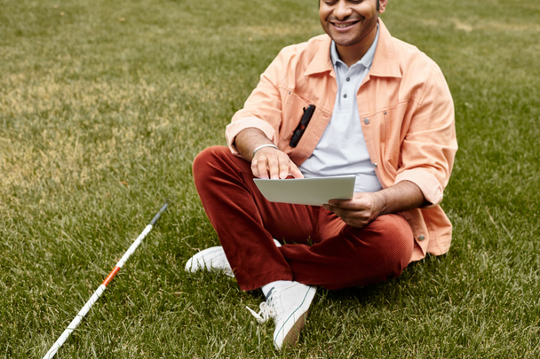 радісний індійський чоловік зі сліпотою в помаранчевій куртці сидить на траві і читає код брайля - Фото, зображення