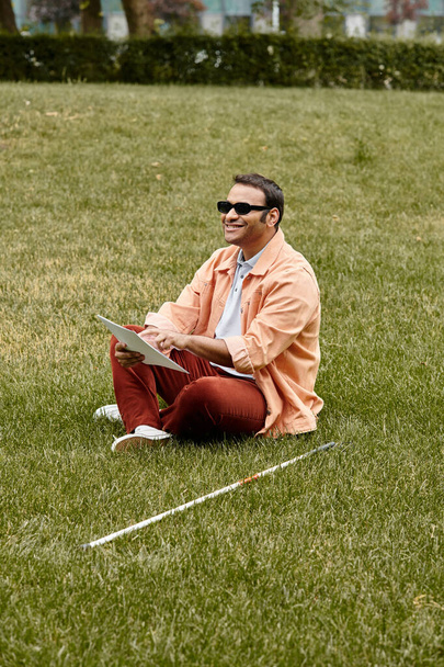 wesoły indiański niewidomy mężczyzna w pomarańczowej żakiecie siedzi na trawie w okularach i czyta kod Braille 'a - Zdjęcie, obraz