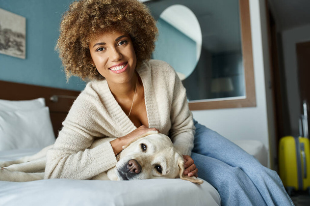 young and happy african american woman cuddling labrador on a bed in a pet-friendly hotel room - Photo, Image