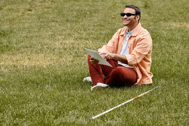 Wesoły indyjski niewidomy mężczyzna w pomarańczowej żakiecie siedzi na trawie w okularach i czyta kod Braille 'a - Zdjęcie, obraz