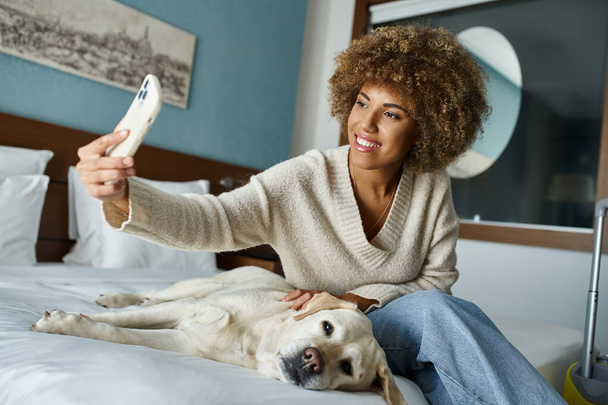 feliz afro-americana tomando selfie com labrador em uma cama em um quarto de hotel pet-friendly - Foto, Imagem