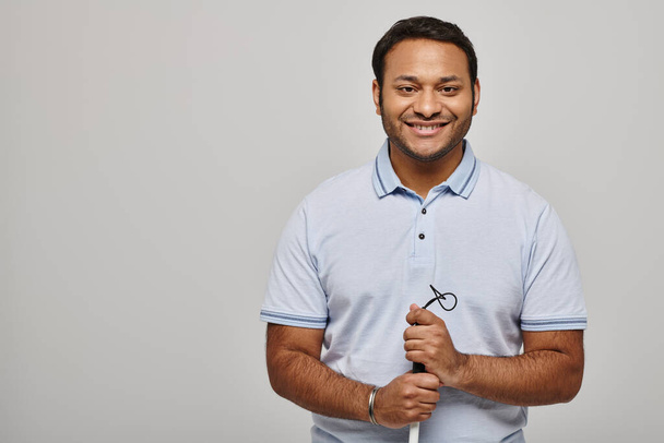 cheerful indian disabled man in blue tee shirt holding walking stick and posing on gray backdrop - Photo, Image