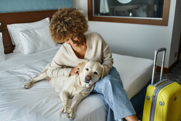 giovane e sorridente donna africana americana coccolando labrador vicino ai bagagli in camera d'albergo pet-friendly - Foto, immagini