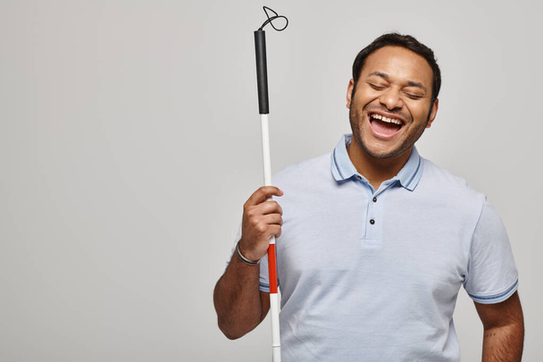 heureux indien handicapé en t-shirt bleu tenant bâton de marche et posant sur fond gris - Photo, image
