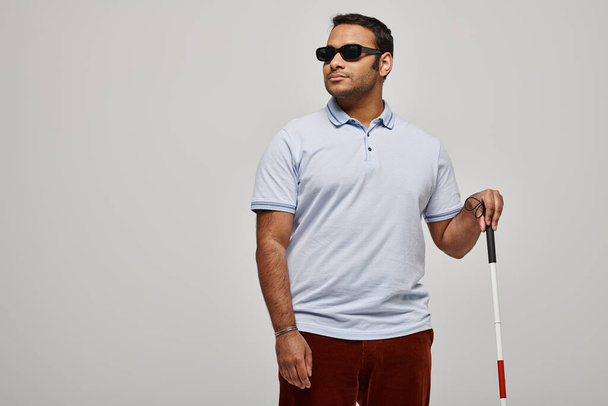 atractivo hombre ciego indio en camiseta azul con gafas y bastón de paseo posando sobre fondo gris - Foto, imagen