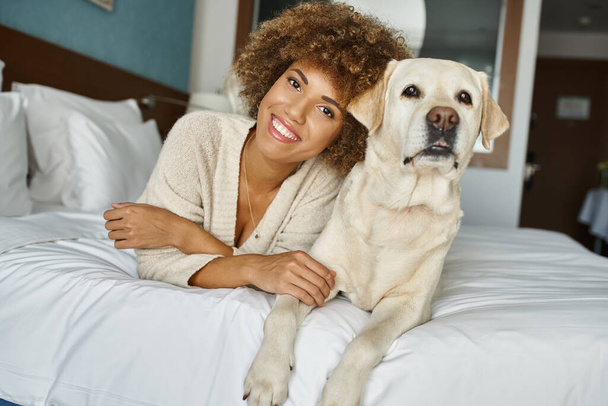 Iloinen afrikkalainen amerikkalainen nainen labradorinsa kanssa makaa sängyssä lemmikkiystävällisessä hotellissa, matkustaa - Valokuva, kuva