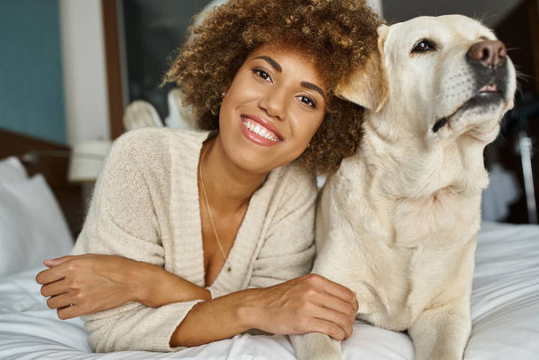 donna afro-americana positiva con il suo labrador sdraiato su un letto in un hotel pet-friendly, viaggi - Foto, immagini