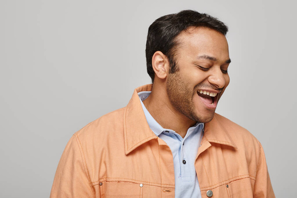 веселый индийский мужчина в яркой оранжевой куртке счастливо улыбается и смотрит в сторону на сером фоне - Фото, изображение