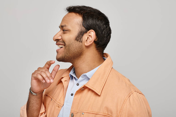 χαρούμενος ινδός άντρας με ζωηρό πορτοκαλί μπουφάν χαμογελώντας χαρούμενα και κοιτάζοντας μακριά σε γκρι φόντο - Φωτογραφία, εικόνα