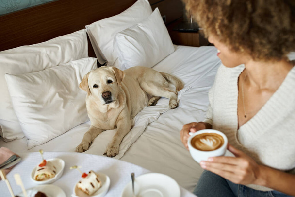afro-amerikai nő kezében cappuccino és nézi labrador kutya állatbarát hotel - Fotó, kép