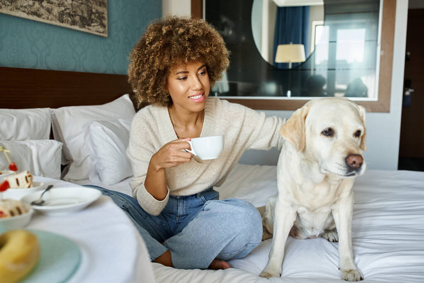 ペットフレンドリーなホテルの部屋でカップとラブラドール犬を抱えている幸せなアフリカ系アメリカ人女性 - 写真・画像