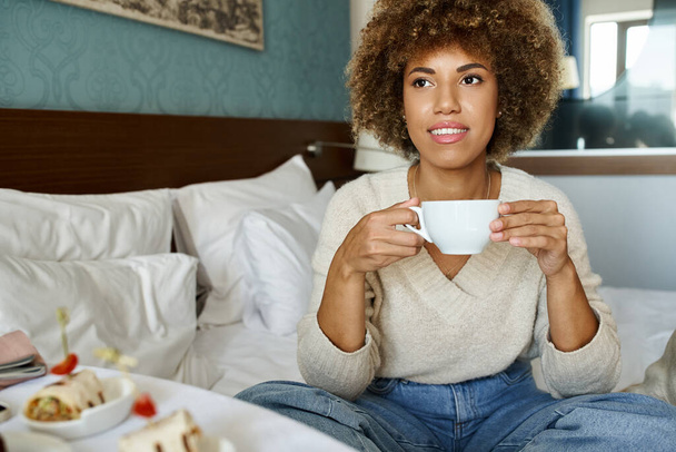 szczęśliwy Afroamerykanka trzyma filiżankę i siedzi na łóżku w pokoju hotelowym, room service i komfort - Zdjęcie, obraz