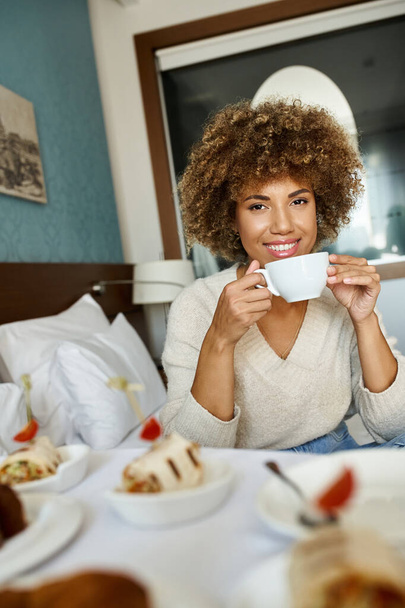 ευτυχισμένη Αφροαμερικανή γυναίκα κρατώντας κύπελλο και κάθεται στο κρεβάτι στο δωμάτιο του ξενοδοχείου, υπηρεσία δωματίου και άνεση - Φωτογραφία, εικόνα