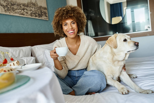 vrolijk Afrikaans amerikaanse vrouw holding cup en knuffelen labrador hond in huisdiervriendelijke hotelkamer - Foto, afbeelding