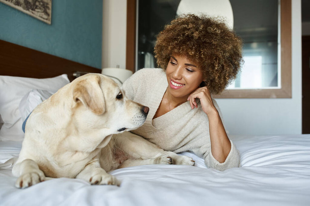 Χαρούμενη Αφροαμερικανή γυναίκα ξαπλωμένη στο κρεβάτι και αγκαλιάζοντας το λαμπραντόρ σκυλί της σε φιλικό προς τα κατοικίδια ξενοδοχείο - Φωτογραφία, εικόνα