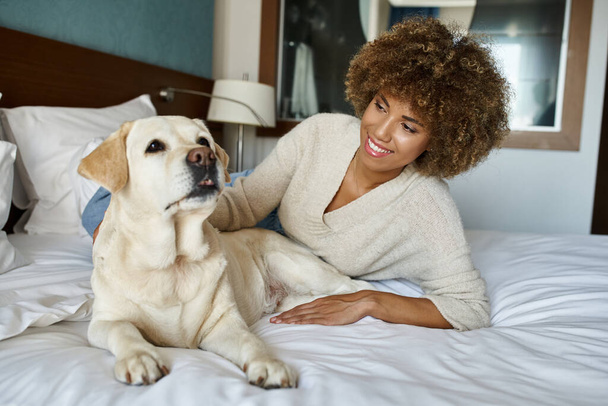 笑顔のアフリカ系アメリカ人女性がベッドに横たわり,ペットフレンドリーなホテルでラブラドール犬を抱きしめる - 写真・画像