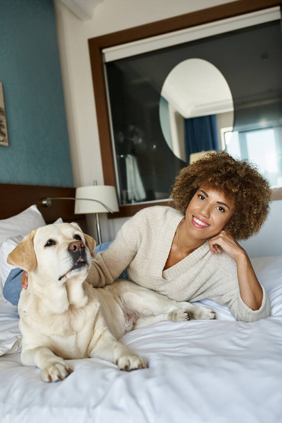 若いアフリカ系アメリカ人女性がベッドに横たわり,ペットフレンドリーなホテルでラブラドール犬を抱きしめる - 写真・画像