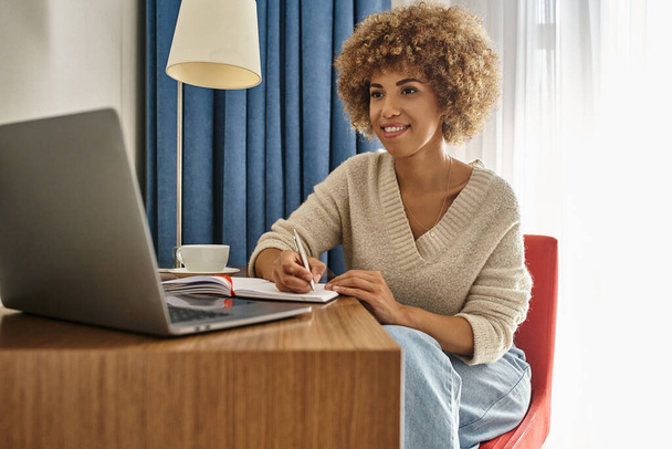 mujer afroamericana feliz a distancia trabajando en la habitación del hotel, usando el ordenador portátil y tomando notas - Foto, Imagen