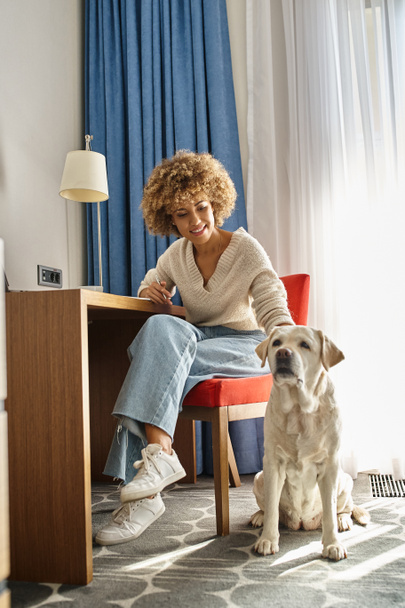 happy african american woman remote working on laptop near her labrador at pet-friendly hotel - 写真・画像