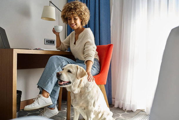 gelukkig Afrikaans amerikaanse vrouw geniet van koffie en werken in de buurt van haar labrador in huisdiervriendelijk hotel - Foto, afbeelding
