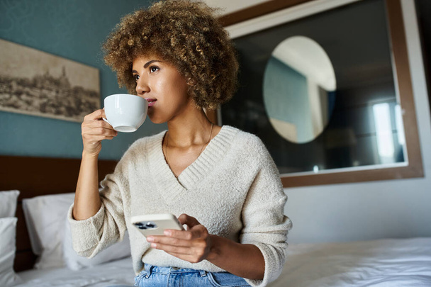 capelli ricci donna afroamericana sorseggiando caffè mentre si utilizza lo smartphone in camera d'albergo, viaggi - Foto, immagini
