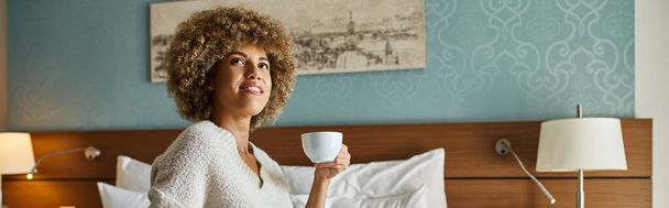 Banner der glücklichen afrikanisch-amerikanischen Frau, die Kaffee genießt, während sie auf dem Hotelbett sitzt, Wochenendausflug - Foto, Bild