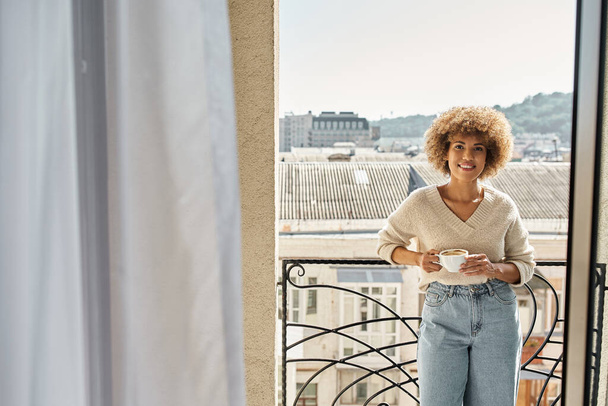 souriante femme afro-américaine frisée debout avec une tasse de café sur le balcon de l'hôtel, vue sur la ville - Photo, image