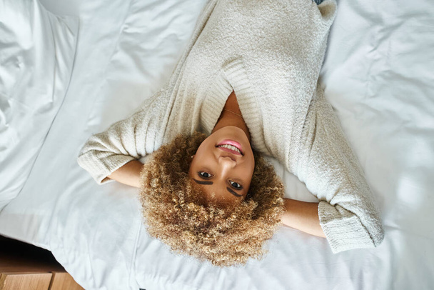 Draufsicht der entspannten afrikanisch-amerikanischen Frau lächelnd und auf dem Bett im Hotelzimmer liegend, verkehrt herum - Foto, Bild