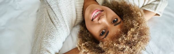 πανό της χαρούμενης Αφροαμερικανής γυναίκας χαμογελώντας και ξαπλωμένη στο κρεβάτι στο δωμάτιο του ξενοδοχείου, ανάποδα - Φωτογραφία, εικόνα
