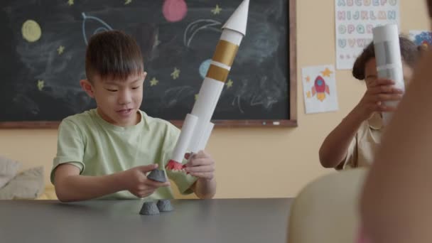 Malý asijský chlapec dělat papírové rakety a hrát si s Afroameričanky, zatímco sedí spolu za stolem ve školní třídě během lekce - Záběry, video