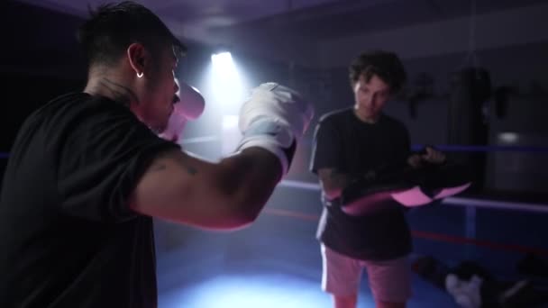Entrenamiento Muay Thai Fighter con la ayuda del soporte de almohadilla. Sportsman perforando y pateando almohadillas dentro del ring de boxeo e iluminación dramática - Metraje, vídeo